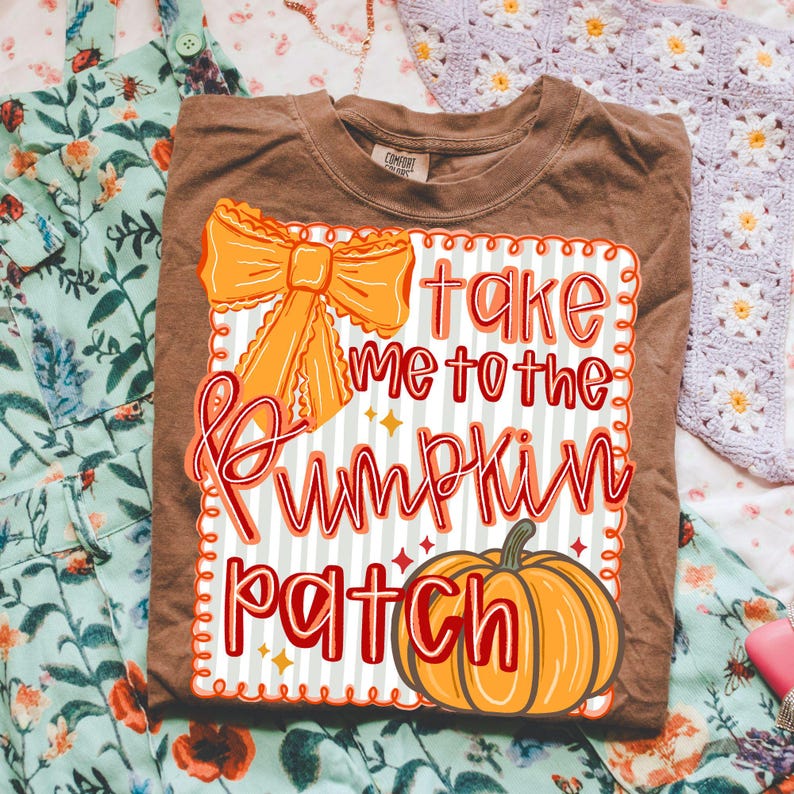 May include: Brown t-shirt with the text "Take Me To The Pumpkin Patch" in red and orange. The design includes an orange bow and a pumpkin illustration. The shirt is folded on a floral patterned surface.