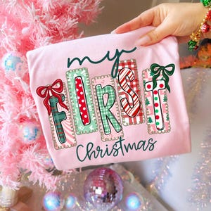 May include: A light pink t-shirt with the text "my FIRST Christmas" in a festive design. The letters are decorated with patterns and bows in red, green, and white, perfect for the holiday season.