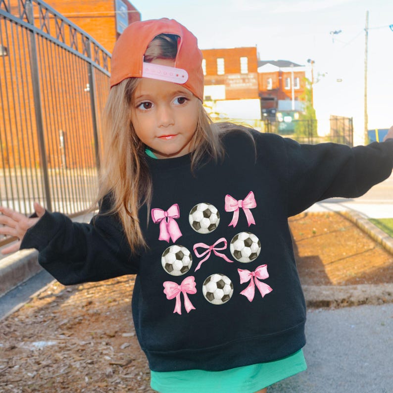 May include: A black sweatshirt featuring a soccer ball and pink bow design. The soccer balls are black and white, and the bows are pink. The sweatshirt is worn by a young person wearing an orange baseball cap.