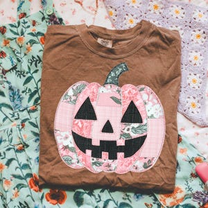 May include: A brown t-shirt featuring a pink and floral patchwork pumpkin design. The pumpkin has black triangle eyes and a jagged mouth. The shirt is folded and rests on a floral patterned fabric.
