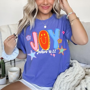 May include: A periwinkle blue t-shirt with the word "JOY" in colorful, outlined letters. The "O" is a smiling face. The text "John 16:22" is below the word "JOY". The shirt is decorated with stars and flowers.