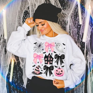May include: White sweatshirt featuring a Halloween-themed design. The design includes pink bows, a haunted house, a ghost, a witch hat, a jack-o'-lantern, and a spider web. Black bats and stars are also included in the design.