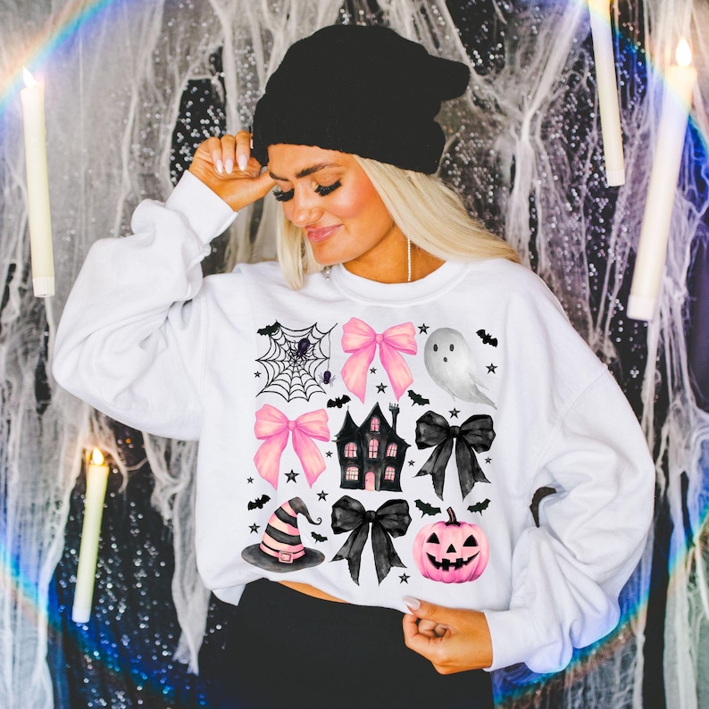 May include: White sweatshirt featuring a Halloween-themed design. The design includes pink bows, a haunted house, a ghost, a witch hat, a jack-o'-lantern, and a spider web. Black bats and stars are also included in the design.