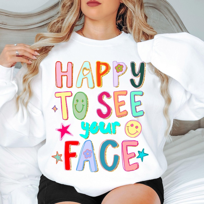 May include: White sweatshirt with the phrase "HAPPY TO SEE YOUR FACE" in colorful, outlined letters. The design includes stars, a heart, a flower, and smiley faces. The sweatshirt has long sleeves.