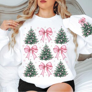 May include: White sweatshirt featuring a festive design with green Christmas trees adorned with pink ornaments and bows. The design is repeated across the front and on the sleeve, with snowflakes scattered around the trees and bows.