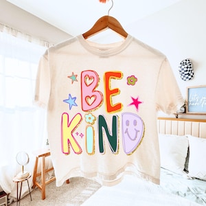 May include: A cream-colored t-shirt with the words "BE KIND" in colorful, playful lettering. The design includes hearts, stars, flowers, and a smiling face. The shirt is hanging on a wooden hanger.