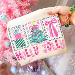 May include: A cream-colored Christmas-themed tote bag with the words "HOLLY JOLLY" in pink. The bag features three framed designs with a gift, Christmas tree, and bow. The designs are on a pink and white plaid background.