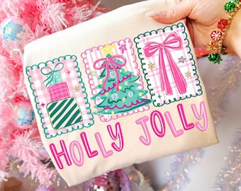 May include: A cream-colored Christmas-themed tote bag with the words "HOLLY JOLLY" in pink. The bag features three framed designs with a gift, Christmas tree, and bow. The designs are on a pink and white plaid background.