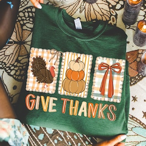 May include: Green sweatshirt with a Thanksgiving theme. Features three square designs with a turkey, pumpkins, and a bow. The words "GIVE THANKS" are printed in a colorful font.