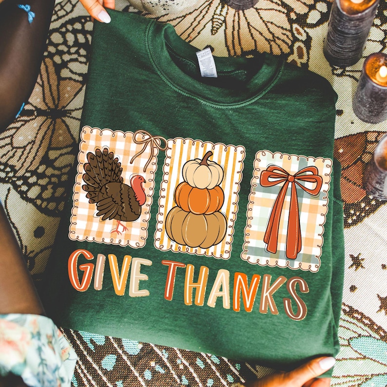 May include: Green sweatshirt with a Thanksgiving theme. Features three square designs with a turkey, pumpkins, and a bow. The words "GIVE THANKS" are printed in a colorful font.