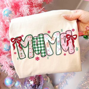 May include: Cream-colored t-shirt with the word "Mama" in a festive design. The letters are decorated with Christmas-themed patterns, including bows, gingerbread men, and snowflakes. The shirt is held up by a hand.
