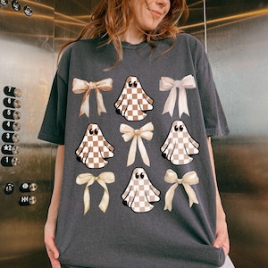 May include: Dark gray t-shirt featuring a pattern of tan and white checkered ghosts and cream-colored bows. The ghosts have quotation marks above them. The shirt is a casual, oversized fit.