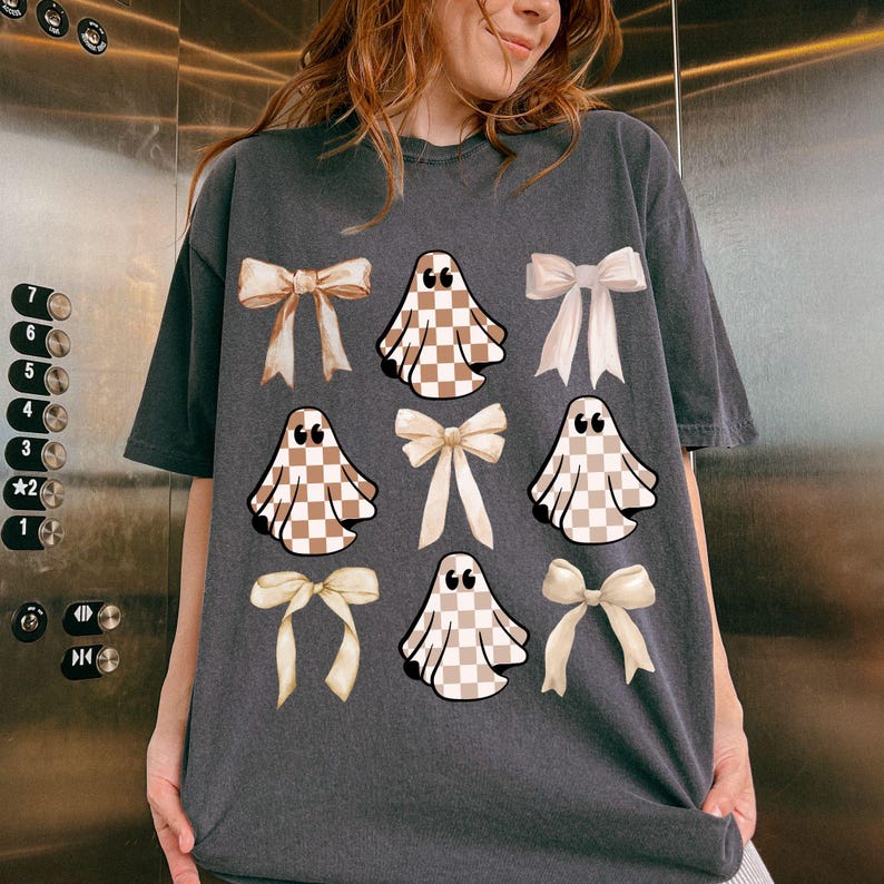 May include: Dark gray t-shirt featuring a pattern of tan and white checkered ghosts and cream-colored bows. The ghosts have quotation marks above them. The shirt is a casual, oversized fit.