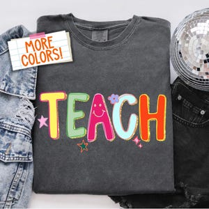 May include: Dark gray t-shirt with the word "TEACH" in colorful, outlined letters. The design includes a smiling face, stars, and a flower. A denim jacket, black jeans, and a disco ball are in the background. The text "MORE COLORS!" is on a sticky note.