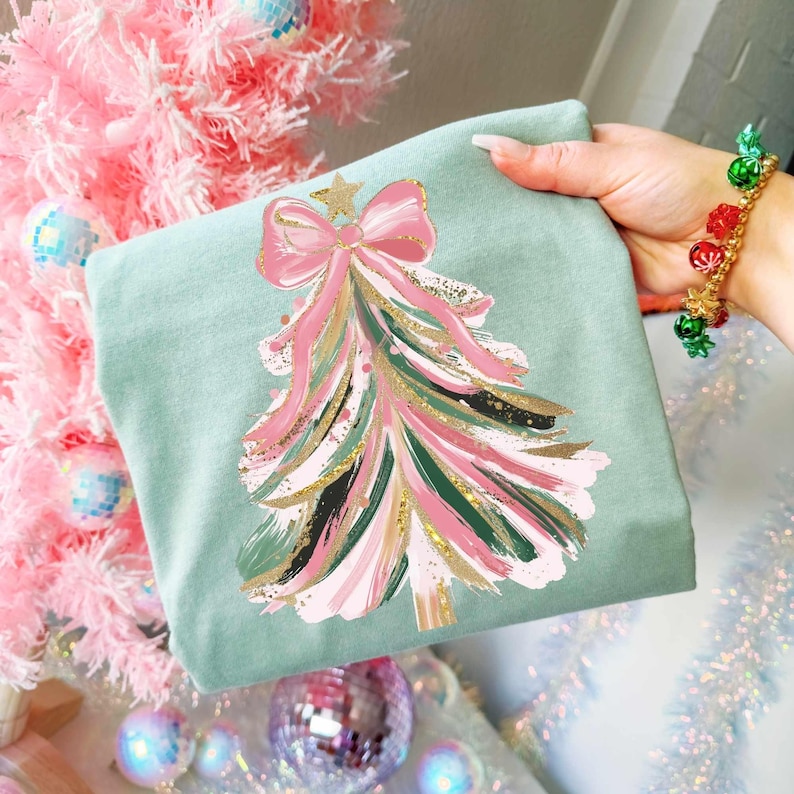 May include: A light green sweatshirt featuring a Christmas tree design. The tree is decorated with pink, green, and gold brushstrokes, topped with a pink bow and a gold star. The sweatshirt is held up by a person.