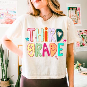 May include: Cream-coloured t-shirt with the words "THIRD GRADE" in colourful, playful lettering. The text features a flower, stars, and a smiley face. The shirt is a casual, comfortable style.