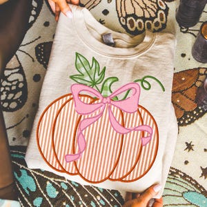 Könnte beinhalten: Beiges Sweatshirt mit einem Kürbismotiv. Der Kürbis hat orange und weiße Streifen, eine rosa Schleife und grüne Blätter. Das Sweatshirt ist gefaltet und wird von Händen gehalten, mit einem gemusterten Hintergrund.
