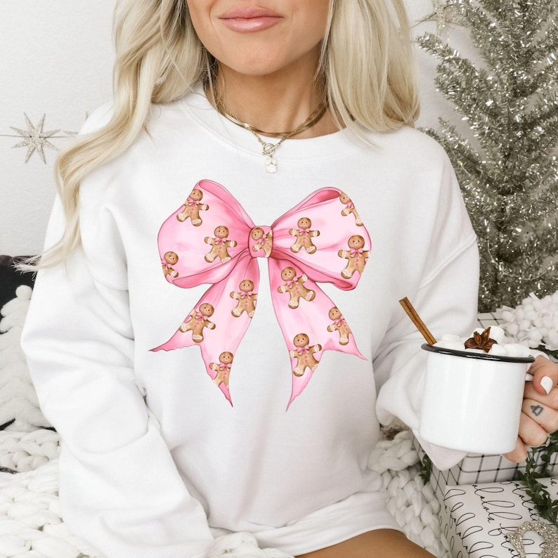 May include: White sweatshirt featuring a large pink bow design adorned with gingerbread men. The bow is the focal point, with the gingerbread men repeating across the bow's surface. The sweatshirt is a festive, holiday-themed garment.