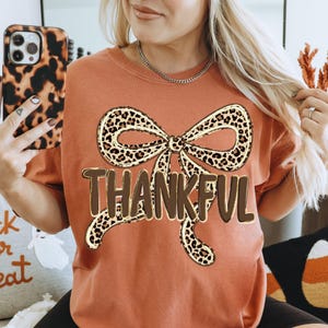 May include: Rust-colored t-shirt featuring a large bow with a leopard print design. The word "THANKFUL" is printed in brown block letters below the bow. The shirt is a casual style, suitable for fall.