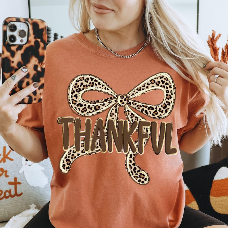May include: Rust-colored t-shirt featuring a large bow with a leopard print design. The word "THANKFUL" is printed in brown block letters below the bow. The shirt is a casual style, suitable for fall.