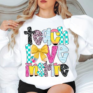 Może przedstawiać: Biała bluza z kolorową grafiką, na której widnieje napis "Teach Love Inspire". Grafika przedstawia kokardkę, ołówki i różne wzory.