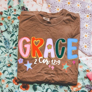 May include: A brown t-shirt with the word "GRACE" in colorful, outlined letters. The design includes a heart, flower, stars, and the text "2 Cor 12:9". The shirt is folded on a floral patterned surface.