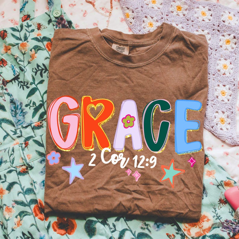 May include: A brown t-shirt with the word "GRACE" in colorful, outlined letters. The design includes a heart, flower, stars, and the text "2 Cor 12:9". The shirt is folded on a floral patterned surface.