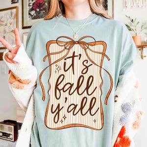 Pode incluir: Camiseta azul clara com a frase "it's fall y'all" em marrom. O texto está dentro de uma moldura marrom com um laço e listras verticais. Uma peça casual para o outono.