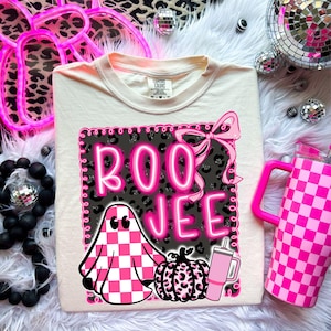 May include: Cream-coloured t-shirt with a pink and black graphic that says "BOO JEE". The graphic includes a checkered ghost, a pumpkin, and a drink cup. A pink bow is at the top of the graphic. A pink checkered tumbler is on the right.