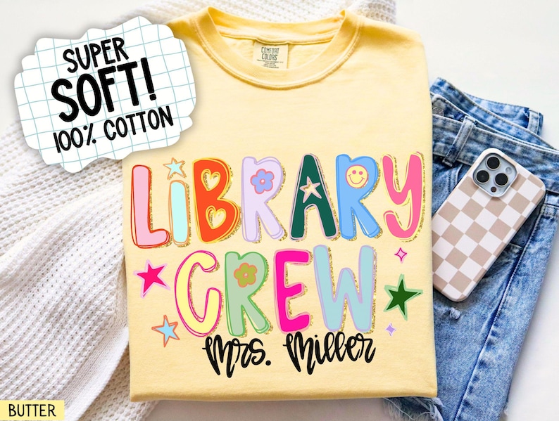 Może przedstawiać: Ż&oacute;łta koszulka z napisem "LIBRARY CREW Mrs. Miller" w kolorowych, zabawnych literach. Koszulka posiada r&oacute;wnież grafiki uśmiechniętych buziek i gwiazdek. Tekst "SUPER SOFT! 100% COTTON" znajduje się w białym dymku.