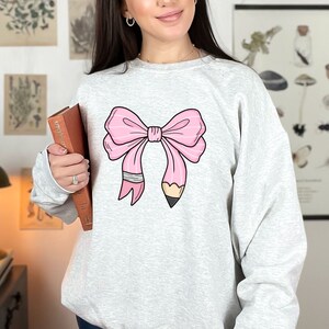Puede incluir: Una sudadera gris con un lazo rosa hecho de un l&aacute;piz. El lazo est&aacute; atado en un nudo y el l&aacute;piz est&aacute; afilado.