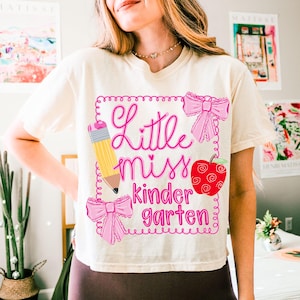 Op de afbeelding: Cr&egrave;mekleurig T-shirt met de tekst "Little Miss Kindergarten" in roze letters. Het ontwerp bevat een gele potlood, roze strikken en een rode appel. Het shirt heeft een casual stijl.