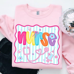 Può includere: Felpa rosa chiaro con la scritta "NURSE" in lettere colorate. Il design include uno stetoscopio, una siringa, pillole e fiori. Lo sfondo presenta strisce blu e bianche.