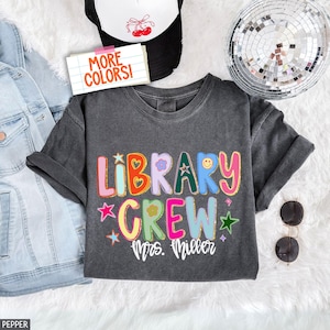 May include: Dark gray t-shirt with the words "LIBRARY CREW Mrs. Miller" in colorful, glittery letters. The shirt has rolled-up sleeves and is surrounded by a denim jacket, a black and white trucker hat, sunglasses, and a disco ball.