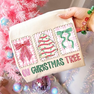 May include: A cream-colored Christmas-themed tote bag featuring three decorative squares with a pink gingham pattern. The squares display a red bow, a Christmas tree, and a green bow. The text "OH CHRISTMAS TREE" is printed below the squares.