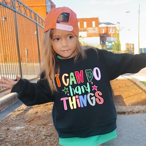 May include: A young person wearing a black sweatshirt with the text "I CAN DO hard THINGS" in colorful letters. The person is also wearing an orange baseball cap with a pink brim and a green skirt.
