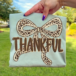 May include: A light blue t-shirt featuring a large bow with a leopard print design. The word "THANKFUL" is printed in brown block letters below the bow. The shirt is held up by a person.