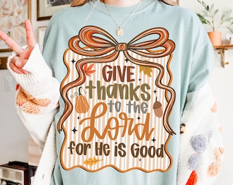 May include: A light blue t-shirt with a fall-themed graphic. The graphic includes a large bow, leaves, a pumpkin, and the text "Give thanks to the Lord for He is Good" in brown and orange.