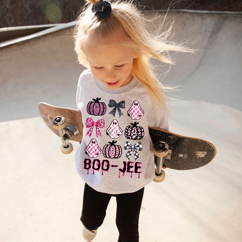 Może przedstawiać: Jasnoszara bluza z motywem Halloween z r&oacute;żowymi, czarnymi i białymi dyniami, duchami i kokardkami. Tekst "BOO-JEE" jest wydrukowany na r&oacute;żowo z kroplowymi akcentami. Dziecko ma na sobie bluzę i trzyma deskorolkę.