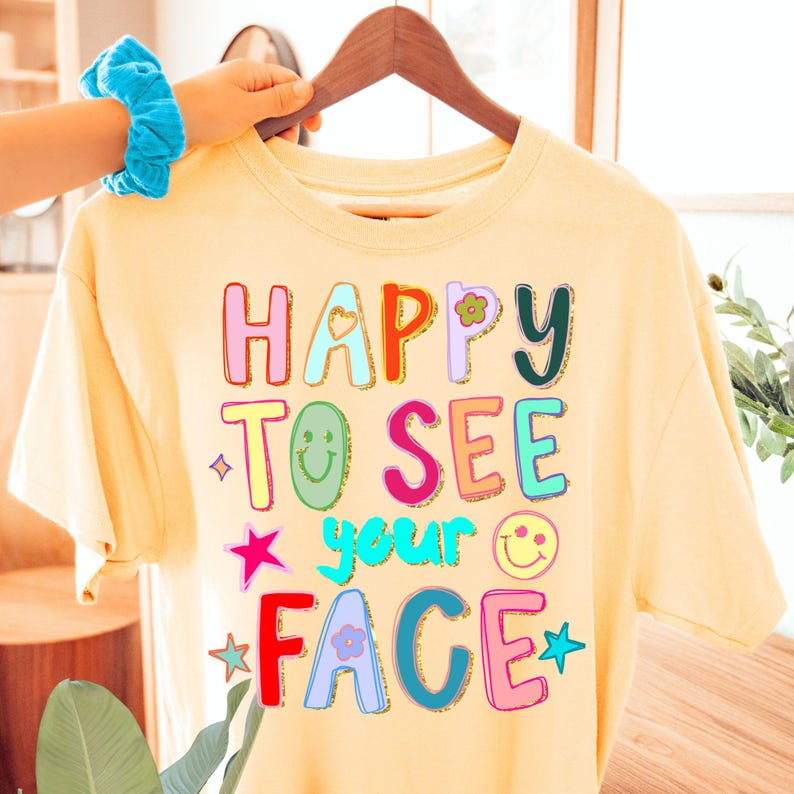 May include: A light yellow t-shirt with the phrase "HAPPY TO SEE your FACE" in colorful, bubble letters. The design includes a heart, flower, star, and smiley face graphics. The shirt is hanging on a wooden hanger.