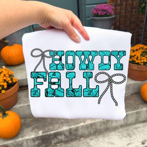 Puede incluir: Sudadera blanca con las palabras "HOWDY FALL" en letras turquesas con un diseño de cuentas negras. La sudadera es sostenida por una mano, con calabazas y flores en macetas en el fondo.