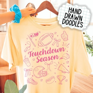 May include: A light yellow t-shirt with a pink graphic design featuring football-themed doodles. The words "Touchdown Season" are prominently displayed. The shirt is held up by a wooden hanger.