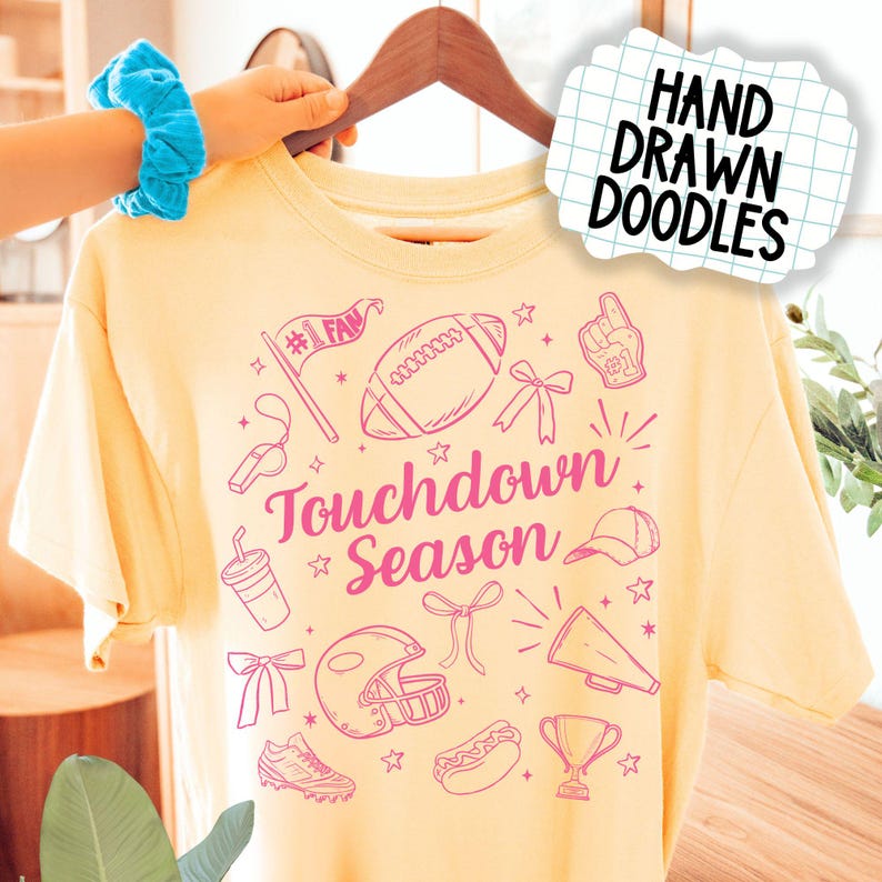 May include: A light yellow t-shirt with a pink graphic design featuring football-themed doodles. The words "Touchdown Season" are prominently displayed. The shirt is held up by a wooden hanger.