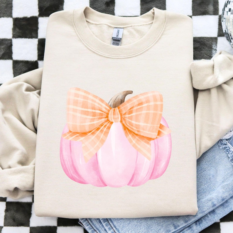 May include: A beige crewneck sweatshirt featuring a watercolor illustration of a pink pumpkin with an orange plaid bow. The pumpkin has a brown stem. The sweatshirt is folded on a black and white checkered surface.