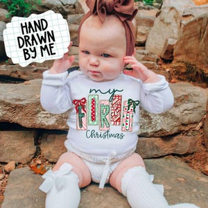 May include: A baby wearing a white long-sleeved shirt with the text "my FIRST Christmas" in a colorful, festive design. The baby is also wearing a brown headband and white socks with bows. The image includes the text "HAND DRAWN BY ME".