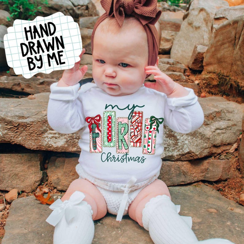 May include: A baby wearing a white long-sleeved shirt with the text "my FIRST Christmas" in a colorful, festive design. The baby is also wearing a brown headband and white socks with bows. The image includes the text "HAND DRAWN BY ME".
