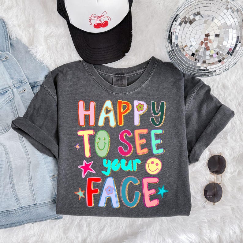May include: A dark gray t-shirt with the words "HAPPY TO SEE your FACE" in colorful, block letters. The shirt is styled with a black trucker hat with a cherry design and sunglasses.