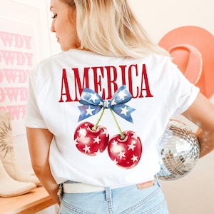 May include: White t-shirt with a red "AMERICA" graphic and a blue and white bow with stars above two red cherries with white stars.