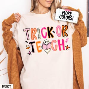 Könnte beinhalten: Elfenbeinfarbenes T-Shirt mit dem Schriftzug "TRICK OR TEACH" in einer farbenfrohen, verspielten Schriftart. Das Design umfasst einen Bleistift, einen Geist, der ein Buch liest, Süßigkeiten und Sterne, ideal für Halloween oder Lehrer.