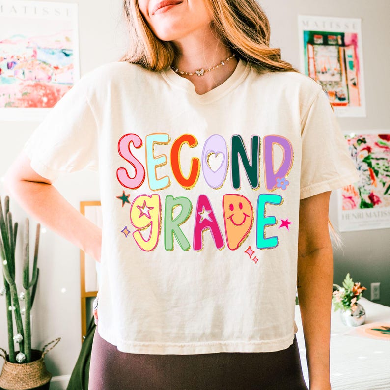 Puede incluir: Camiseta color crema con las palabras "SECOND GRADE" en letras coloridas y de burbujas. El dise&ntilde;o incluye estrellas, un coraz&oacute;n y una carita sonriente. Es una prenda casual para el d&iacute;a a d&iacute;a.